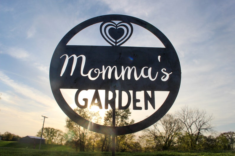 May include: A black metal sign with a circle shape and a heart cut out in the center. The sign says "Momma's Garden" in white lettering.
