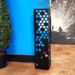 May include: A tall, black metal decorative panel with a honeycomb pattern cut out, revealing a blue interior. The panel stands upright on a brown floor, with a blue wall and a green plant in the background.