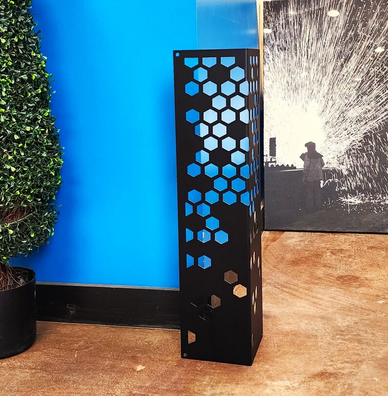 May include: A tall, black metal decorative panel with a honeycomb pattern cut out, revealing a blue interior. The panel stands upright on a brown floor, with a blue wall and a green plant in the background.