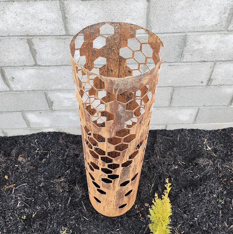 May include: A rusty metal planter with a hexagonal pattern cut out. The planter is standing on a bed of dark brown mulch.