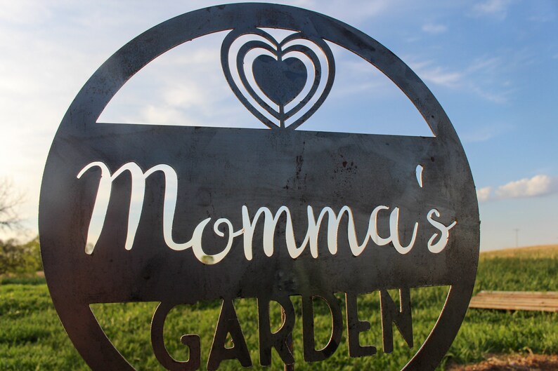 May include: A metal sign with a heart cut out in the top and the words "Momma's Garden" in white lettering.