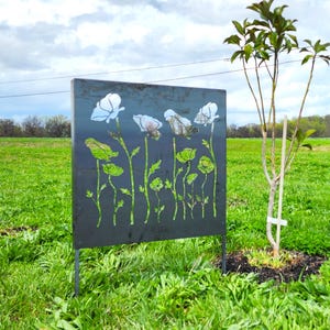 May include: A black metal garden screen with a cut-out design of white flowers. The screen is standing in a grassy field with a small tree in the background.