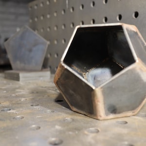 May include: A metal planter with a hexagonal shape, made of welded metal, with a rough, industrial finish. The planter is sitting on a metal surface with a pattern of holes.