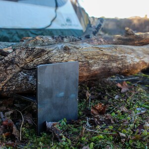 May include: A set of two metal fire grate stands, made of steel, with a rustic log resting on top. The stands are in a grassy area with fallen leaves.