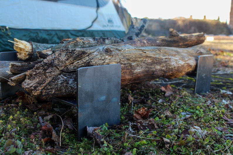 May include: A set of two metal fire grate stands, made of steel, with a rustic log resting on top. The stands are in a grassy area with fallen leaves.