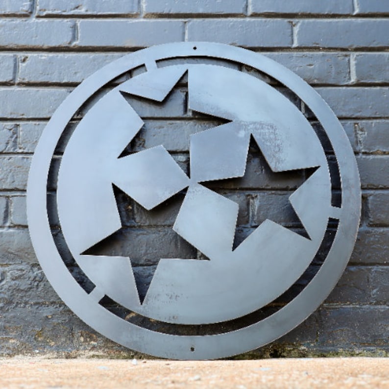 K&ouml;nnte beinhalten: Eine runde Wanddekoration aus Metall mit einem ausgeschnittenen Design aus drei Sternen. Die Sterne sind in einem Kreis angeordnet, wobei die Spitzen der Sterne nach au&szlig;en zeigen. Das Metall ist dunkelgrau.