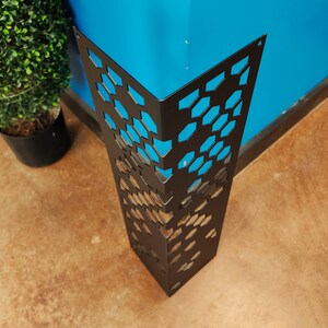 May include: A black metal corner protector with a geometric honeycomb pattern. The protector is designed to fit around a corner, with a blue wall visible through the cutouts. The floor is a light brown color.