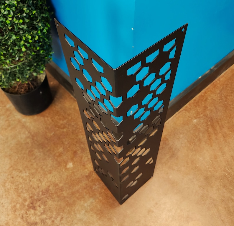 May include: A black metal corner protector with a geometric honeycomb pattern. The protector is designed to fit around a corner, with a blue wall visible through the cutouts. The floor is a light brown color.
