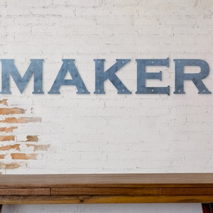 Könnte beinhalten: Ein rustikaler Holztisch mit einer weißen Backsteinwand im Hintergrund. Das Wort "MAKER" ist in großen, blauen Metallbuchstaben an der Wand geschrieben.