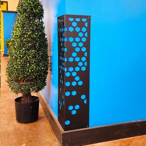 May include: A black metal decorative panel with a honeycomb pattern against a bright blue wall. The panel is rectangular and stands upright. A potted topiary is to the left. The panel is likely a decorative element or a functional cover.