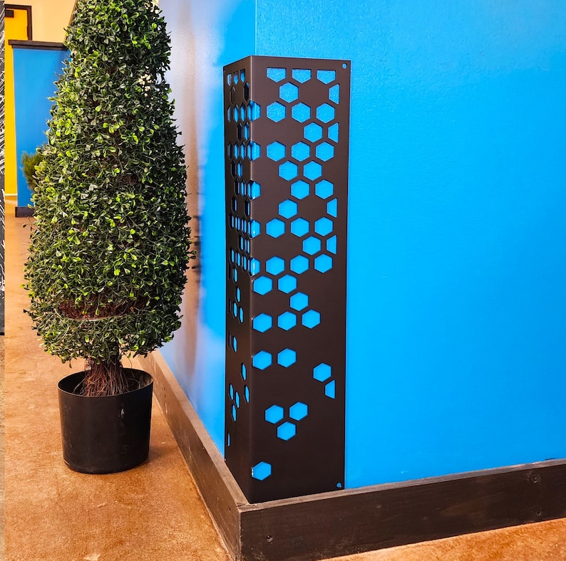 May include: A black metal decorative panel with a honeycomb pattern against a bright blue wall. The panel is rectangular and stands upright. A potted topiary is to the left. The panel is likely a decorative element or a functional cover.