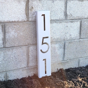 May include: White metal house number sign with the numbers 151 cut out. The sign is mounted on a brick wall.