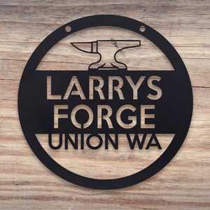 May include: Black metal sign with a circular design. The sign features a silhouette of an anvil and hammer with the text "Larry's Forge Union WA".