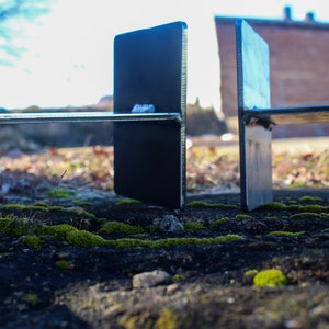 May include: Two black metal brackets with a horizontal metal bar connecting them. The brackets are standing on a mossy surface.
