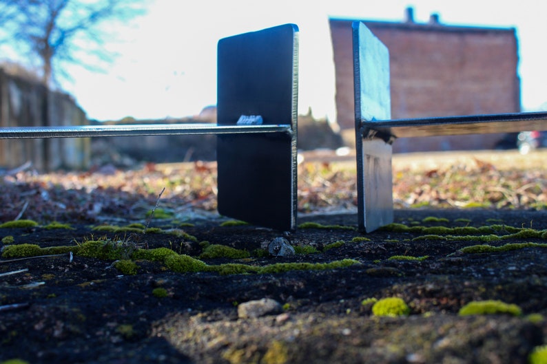 May include: Two black metal brackets with a horizontal metal bar connecting them. The brackets are standing on a mossy surface.