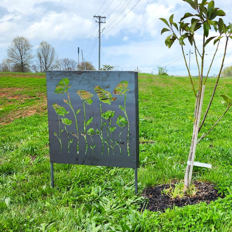 May include: A metal garden art piece with a cutout design of flowers and leaves. The art is standing in a grassy field with a small tree in the background.