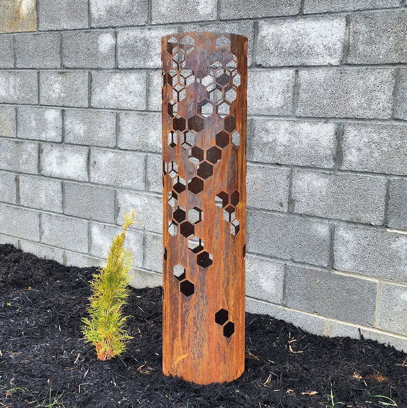May include: A rusty metal planter with a hexagonal pattern cut out. The planter is standing on a bed of black mulch.