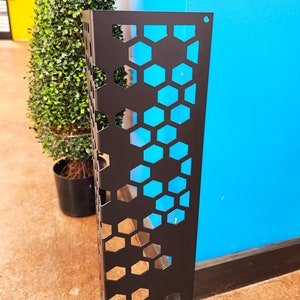 May include: A black metal decorative panel with a honeycomb pattern. The panel is rectangular and has a series of hexagonal cutouts. A green plant in a black pot is visible in the background, along with a blue wall.