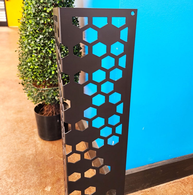 May include: A black metal decorative panel with a honeycomb pattern. The panel is rectangular and has a series of hexagonal cutouts. A green plant in a black pot is visible in the background, along with a blue wall.
