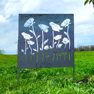 May include: A metal garden decoration with a silhouette of white flowers cut out of the metal. The decoration is standing in a grassy field.