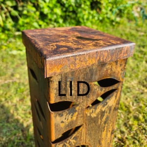 May include: A weathered, square metal container with a lid, featuring leaf-shaped cutouts. The metal has a rusty, orange-brown patina, and the word "LID" is printed on the side. The design suggests outdoor use.