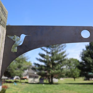 May include: A metal bracket with a hummingbird cutout design. The bracket is dark gray and has a curved shape with a circular hole at the end. It is mounted on a wooden post, with a blue sky and trees in the background.