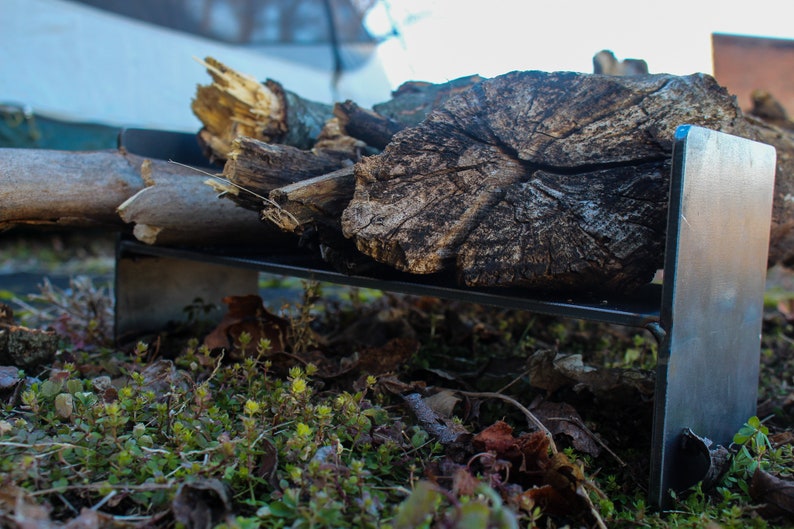 May include: A black metal stand with a piece of firewood resting on top. The stand is sitting on a bed of green moss and brown leaves.