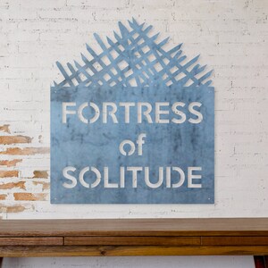 May include: A blue metal sign with a white distressed font that reads "Fortress of Solitude". The sign is mounted on a white brick wall with a red brick accent wall to the left. A wooden table sits in front of the sign.