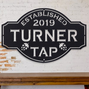 May include: Black metal sign with white text that reads "Established 2019 Turner Tap". The sign has two stylized hop cones on either side of the text "Tap".