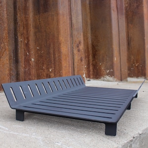 May include: Black metal fireplace grate with a rectangular shape and a series of horizontal bars. The grate has a slight angle and is sitting on a concrete surface.