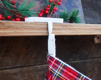Puede incluir: Un gancho de metal blanco para colgar calcetines navideños se apoya en el borde de un estante de madera, con un lazo de tela blanca unido a un calcetín de cuadros rojos, verdes y amarillos que cuelga debajo del estante decorado con pino.
