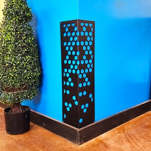Peut inclure: Un pilier décoratif en métal brun foncé avec un motif en nid d'abeille découpé, révélant un intérieur bleu vif. Le pilier se dresse dans un coin contre un mur bleu, à côté d'un petit arbre topiaire vert dans un pot noir.