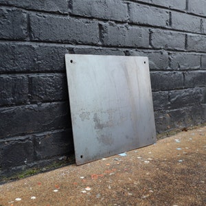 K&ouml;nnte beinhalten: Ein schlichtes, rechteckiges Metallschild mit vier L&ouml;chern an den Ecken. Das Schild lehnt an einer schwarzen Backsteinmauer.