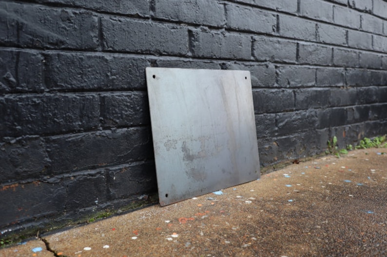 K&ouml;nnte beinhalten: Ein schlichtes, rechteckiges Metallschild mit vier L&ouml;chern an den Ecken. Das Schild lehnt an einer schwarzen Backsteinmauer.