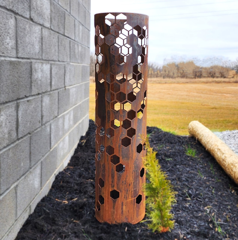 May include: A rusty metal cylinder with a hexagonal pattern cut out. The cylinder is standing upright on a bed of black mulch.