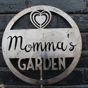 May include: A metal sign with a heart design and the text "Momma's Garden" in a circular shape.