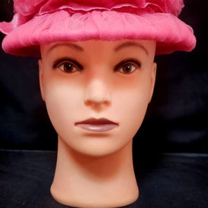 Puede incluir: Un sombrero rosa con ala ancha y una gran corona de tul con volantes. El sombrero se exhibe sobre una cabeza de maniquí con tono de piel neutro, sobre un fondo negro. El color del sombrero es un rosa vibrante y llamativo.