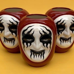 Puede incluir: Tres tazas de cerámica roja con diseños de cara en blanco y negro. Las caras tienen ojos negros, llamas negras alrededor de la cara y una boca blanca con dientes negros.