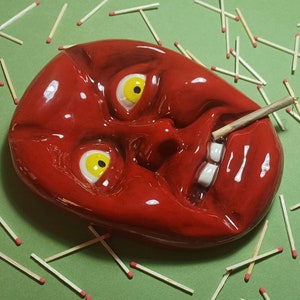 Può includere: Un posacenere in ceramica rossa a forma di viso con tratti esagerati, tra cui occhi grandi, una bocca larga e denti. Un solo fiammifero è posto nella bocca del posacenere.