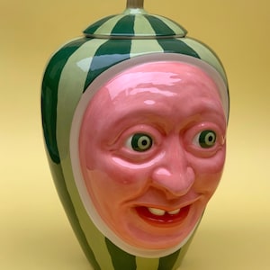 Könnte beinhalten: Eine Keramikvase in Form einer Wassermelone mit einem grün-weiß gestreiften Muster und einem menschlichen Gesicht mit rosa Haut, grünen Augen und einem breiten Lächeln.