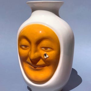 Pode incluir: Um vaso de cerâmica branco com um design de rosto sorridente em tons de laranja e amarelo. Uma pequena mosca está pousada no rosto. O vaso tem um acabamento liso e brilhante e uma forma arredondada, com um pescoço estreito.
