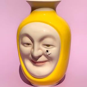 Könnte beinhalten: Eine gelbe Keramikvase mit einem weißen Gesicht und einer Fliege auf der Nase. Die Vase hat eine abgerundete Form und eine kleine Öffnung oben.