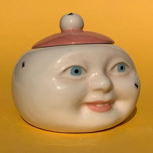 Può includere: Un barattolo di ceramica bianca con un design a forma di viso sorridente, occhi blu e un coperchio rosa. Il barattolo ha una forma rotonda e piccoli dettagli di insetti neri. Il coperchio ha un piccolo pomello bianco.