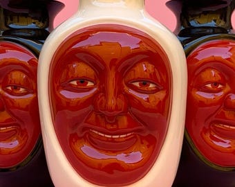 May include: Three ceramic vases with a red face design. The center vase is white with a red face, while the vases on either side are black with a green face.