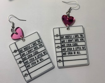 May include: A pair of earrings with a pink heart charm and a white paper notepad with the text "BUT MOSTLY I HATE THE WAY I DON'T HATE YOU. NOT EVEN CLOSE. NOT EVEN A LITTLE BIT. NOT EVEN AT ALL."