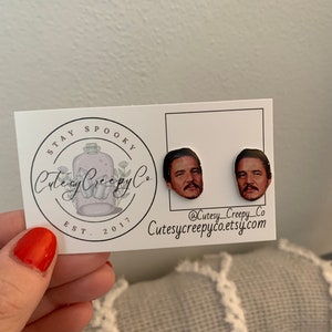 May include: A pair of earrings featuring a portrait of a man with a mustache and beard. The earrings are on a white card with the text "Stay Spooky CutesyCreepyCo Est. 2017 @Cutesy_Creepy_Co Cutesycreepyco.etsy.com"