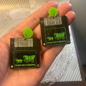 May include: A pair of black and green earrings shaped like floppy disks with a green wagon and the text "You have died of dysentery" on each disk. The earrings are hanging from green circular charms.