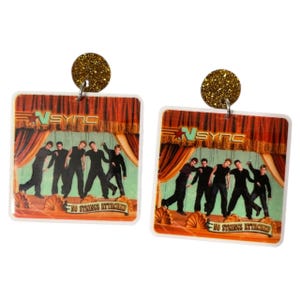 May include: A pair of square earrings featuring an image of the band *NSYNC on a stage. The earrings have a gold glitter top and the image has the text "NO STRINGS ATTACHED".