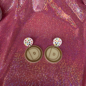 May include: A pair of white wooden earrings with a colorful confetti design and a brown wooden circle with the letter "D" in the center.