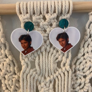May include: Heart-shaped earrings featuring a portrait of a person. The earrings have a white background with a photo of a person in a brown shirt. The earrings are attached to a teal green circular stud.